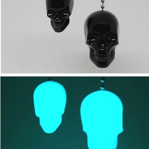May include: Two black skull-shaped light pulls hanging from a chain. The skulls have detailed facial features. The bottom image shows the same skulls glowing with a bright blue light. The background is a solid color.
