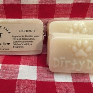 May include: Two bars of Dirty Dog Soap on a red and white checkered cloth. One bar has a label with a goat logo and text. The other bar has a paw print design and the words "Dirty Dog".