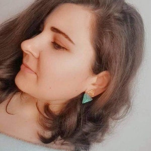 May include: A woman with long brown hair wearing a light blue fuzzy sweater and a pair of green and gold triangle earrings.