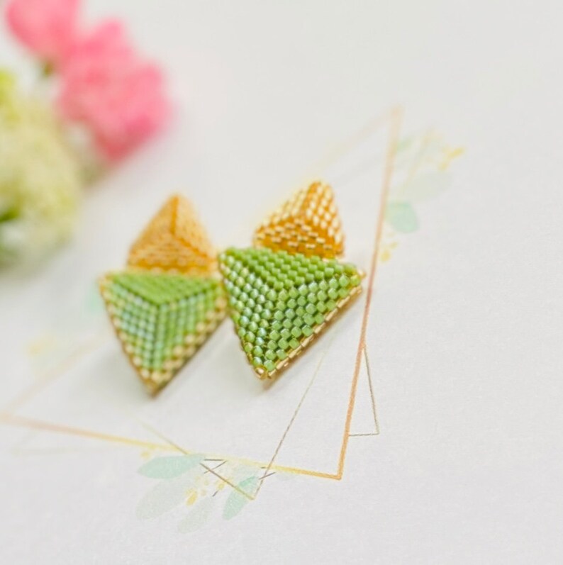 May include: A pair of green and gold triangle-shaped earrings made of beads. The earrings are small and delicate.