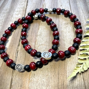 Può includere: Due braccialetti con perline alternate nere e rosse. Ogni braccialetto ha un ciondolo d'argento con un simbolo triangolare.