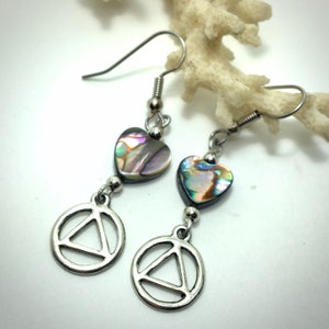 Puede incluir: Un par de pendientes de plata con un dije de concha de abulón en forma de corazón y un dije de círculo plateado con un triángulo en su interior.