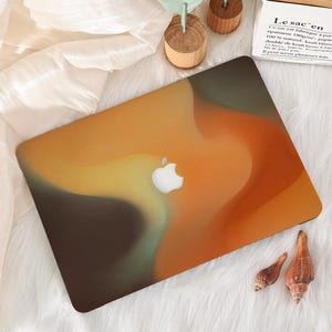 May include: A laptop with a colorful abstract design in shades of orange, yellow, and brown. The Apple logo is visible on the laptop's surface. The laptop is on a white, fluffy surface.