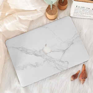 May include: A white laptop featuring a marble effect design and the Apple logo. The laptop is placed on a white, fluffy surface, with wooden decorations and seashells. The style is clean and minimalist.