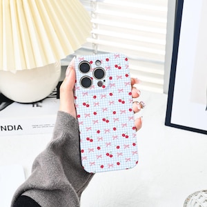 Op de afbeelding: Een witte telefoonhoes met een patroon van roze strikken en rode kersen op een lichtblauwe en witte geruite achtergrond.