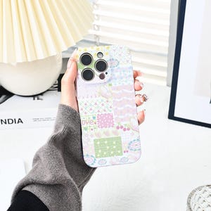 Puede incluir: Una funda de teléfono de color pastel con un diseño de patchwork. La funda presenta cuadrados con patrones de cerezas, mariposas, corazones y estrellas. La palabra "love" está impresa en la funda. La funda está diseñada para un teléfono inteligente.