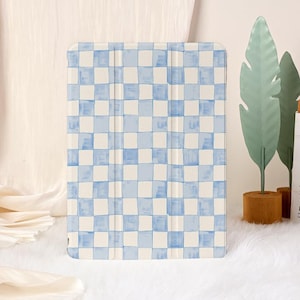 May include: A tablet case featuring a blue and white chequered design. The case is rectangular with a folding cover. The pattern resembles a watercolour painting. The case is displayed on a white, fluffy surface.