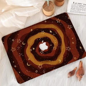 May include: A laptop skin featuring a swirling vortex pattern in shades of dark brown, rust, and golden yellow. The design is accented with gold-colored outlines of stars and crescent moons scattered across the surface, centered around the Apple logo.