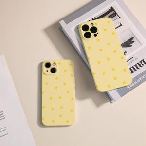 Puede incluir: Dos fundas amarillas para teléfono con un patrón repetido de pequeñas estrellas amarillas. Las fundas tienen recortes para la cámara y los botones. Una funda está plana, la otra está sobre un libro.