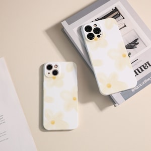 May include: Two white iPhone cases with a floral pattern and a yellow tint. The cases have a camera lens cutout and are on a white surface.
