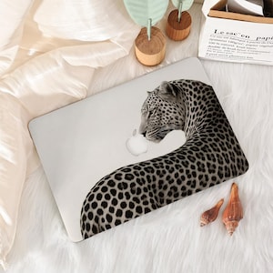 May include: A laptop with a leopard print design. The black and white leopard pattern covers the back of the laptop, with the leopard's head and neck visible. The Apple logo is visible on the laptop. The laptop is on a white, fluffy surface.
