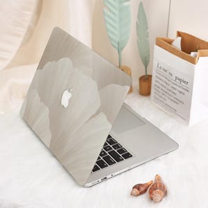 May include: A silver laptop with a light beige floral design on the lid. The design features large, abstract flower shapes. The laptop is open, revealing the keyboard. The background includes decorative elements and a paper bag.