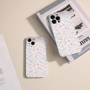 Op de afbeelding: Twee witte iPhone-hoesjes met een bloemenpatroon. De hoesjes hebben een witte achtergrond met kleine roze, blauwe en witte bloemen. De hoesjes hebben een uitsparing voor de camera.