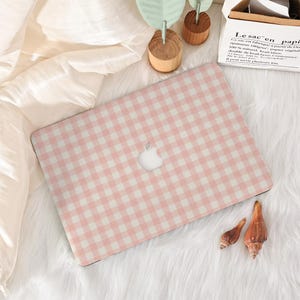 May include: A laptop skin featuring a light pink and white gingham checkered pattern. The rectangular skin is applied to the closed lid of a laptop, displaying a centered silver apple logo cutout. The design consists of a repeating grid.