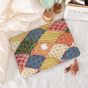 May include: A laptop case with a patchwork design in various colours. The case features red plaid, yellow stripes, and blue floral prints. The Apple logo is visible. The case is on a white, fluffy surface.