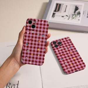 Op de afbeelding: Twee telefoonhoesjes met een rood-roze ruitpatroon en kleurrijke steraccenten. Eén hoesje wordt in de hand gehouden, de andere ligt plat. De hoesjes hebben camera-uitsparingen.
