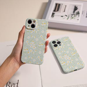 May include: Two light green phone cases with a daisy floral pattern. One case is held in a hand, while the other lies flat. The cases have a soft, matte finish and are designed to fit a smartphone.