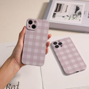 May include: Two lavender phone cases featuring a gingham design. One is held in a hand, the other lies flat. The cases have camera cutouts and a smooth finish. The cases are designed to protect the phone. The cases are a soft shade of purple.