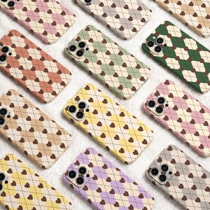 May include: A collection of smartphone cases featuring a repeating argyle pattern with small brown hearts at each intersection. The cases are available in various pastel and solid colours including pink, green, yellow, purple, and beige.