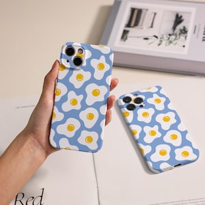 May include: Two phone cases featuring a repeating pattern of fried eggs on a light blue background. One case is held in a hand, the other lies flat. The eggs have white egg whites and yellow yolks.