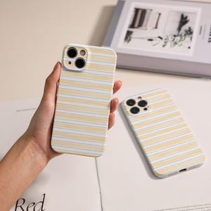 Könnte beinhalten: Zwei Handyhüllen mit gelb-weißem Streifenmuster. Eine Hülle wird in der Hand gehalten, die andere liegt flach. Die Hüllen haben einen Kameraausschnitt und sind für ein Smartphone konzipiert.