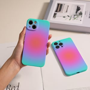 May include: Two phone cases with a gradient design. The cases transition from teal to purple, with a bright pink center. One case is held in a hand, while the other lies flat. The cases have a camera cutout.