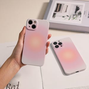 May include: Two pink and white phone cases with a gradient design. One case is held in a hand, the other is lying on a white surface. The cases have a camera cutout and a soft, blurred gradient effect.
