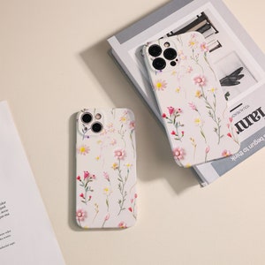 May include: Two white iPhone cases with a floral pattern. The cases have a camera lens cutout and a white background with pink and white flowers.
