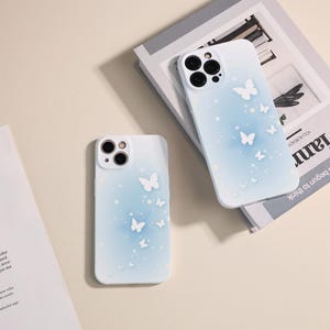 May include: Two white phone cases with a blue gradient and white butterfly designs. The cases have a clear camera lens cutout.