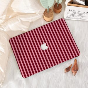 Op de afbeelding: Een laptophoesje met een verticaal gestreept patroon in rood- en roze tinten. Het witte Apple-logo is zichtbaar. De hoes ligt op een wit, getextureerd oppervlak met decoratieve elementen.
