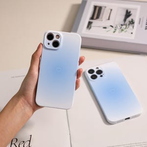 Może przedstawiać: Dwa jasnoniebieskie etui na telefon z motywem serca. Jedno etui jest trzymane w dłoni, drugie leży na białej powierzchni. Etui mają efekt gradientu, przechodząc od bieli do błękitu. Etui mają wycięcie na aparat i port ładowania.