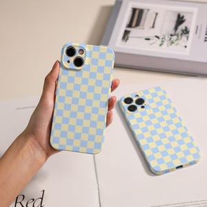 May include: Two light blue and yellow checkered phone cases. One case is held in a hand, while the other rests on a white surface. The cases have a square pattern design and are likely made of a protective material.