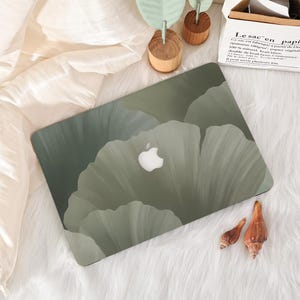 May include: A laptop case featuring a floral design in shades of green and gray. The Apple logo is visible. The case is on a white, fluffy surface, with seashells and wooden decor nearby.