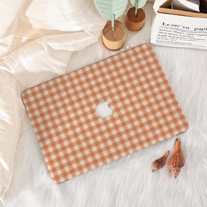 May include: A laptop computer featuring a light orange and white gingham checkered pattern on its exterior lid. The device displays a centered silver apple logo. The laptop is positioned on a white textured surface next to two small seashells.