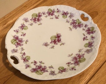 Delicate, handled, handpainted collector plate. It is just over 9 1/2 inches wide. With small lavender and purple flowers.