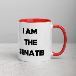 Peut inclure: Une tasse à café en céramique blanche avec un bord et une poignée rouges. La tasse porte une inscription noire qui dit "I AM THE SENATE !"