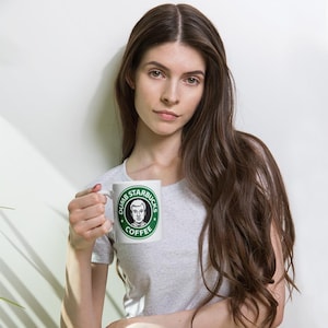 Pode incluir: Caneca de café de cerâmica branca com o texto "Dumb Starbucks Coffee" em um logotipo verde e branco. A caneca apresenta um retrato de um homem no centro do logotipo.