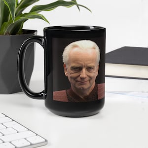 May include: Black ceramic mug with a portrait of a man with white hair and a red and brown outfit. The mug has a black handle. The image is a close-up of the man's face. The mug is a standard size.