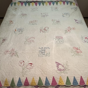 May include: A white quilt with a scalloped edge and a colorful triangle border. The quilt is embroidered with various floral designs in shades of pink, red, green, and blue.