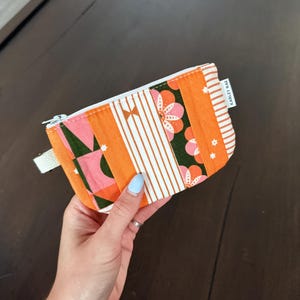 May include: A rectangular fabric zipper pouch featuring a patchwork design of orange, pink, and dark green panels. The patterns include white stripes, small stars, and floral motifs. A white tag with the text KARLEY RAE is attached to the side.