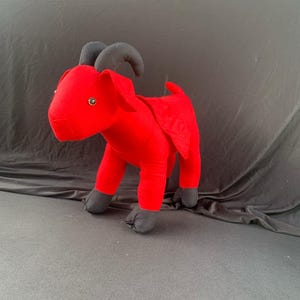 May include: A red plush toy goat with black horns, hooves, and eyes. The goat is standing on a black surface.