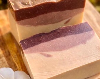 May include: Two handmade soap bars with layered colors of brown, purple, and cream. The top layer of one bar has a swirled design and small gold-colored embellishments. A white flower with a yellow center is in the foreground.