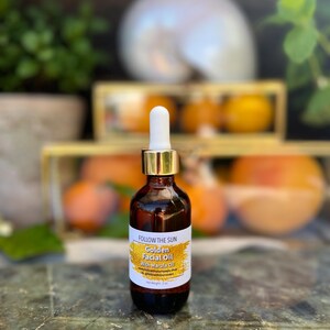 May include: A glass bottle of Golden Facial Oil with Marula Oil by Follow the Sun. The bottle is amber colored with a gold lid and a white dropper. The bottle is sitting on a textured surface.