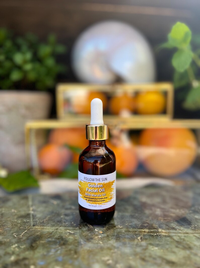 May include: A glass bottle of Golden Facial Oil with Marula Oil by Follow the Sun. The bottle is amber colored with a gold lid and a white dropper. The bottle is sitting on a textured surface.