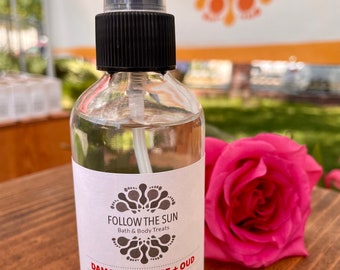 May include: A clear glass bottle with a black spray nozzle and a white label. The label reads "Follow the Sun Bath & Body Treats Damascus Rose + Oud Room & Linen Spray 4oz (118ml) www.followthesunsoap.com IG: @followthesunsoap". A pink rose is in the background.
