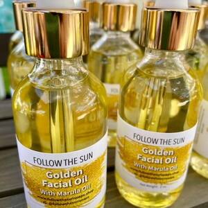 May include: Four amber glass bottles with gold lids and droppers filled with a golden liquid. Each bottle has a white label with the text "FOLLOW THE SUN" and "Golden Facial Oil with Marula Oil". The label also includes the website and Instagram handle for the brand. The bottles are sitting on a wooden surface.
