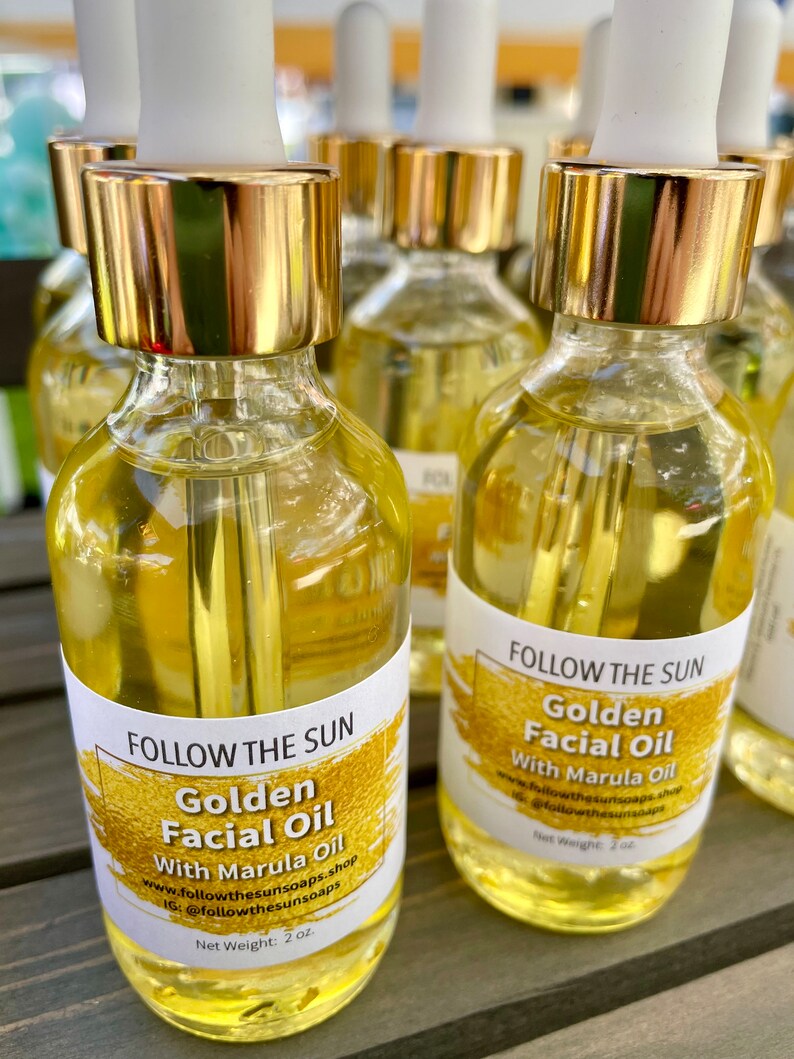 May include: Four amber glass bottles with gold lids and droppers filled with a golden liquid. Each bottle has a white label with the text "FOLLOW THE SUN" and "Golden Facial Oil with Marula Oil". The label also includes the website and Instagram handle for the brand. The bottles are sitting on a wooden surface.