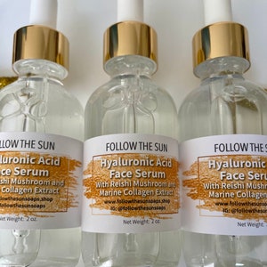 May include: Three glass bottles with gold lids and droppers filled with clear liquid. Each bottle has a white label with the text "FOLLOW THE SUN Hyaluronic Acid Face Serum" and details about the ingredients and where to find the product. The bottles are labeled with a net weight of 2 oz.