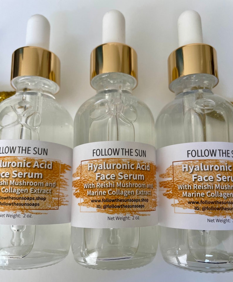 May include: Three glass bottles with gold lids and droppers filled with clear liquid. Each bottle has a white label with the text "FOLLOW THE SUN Hyaluronic Acid Face Serum" and details about the ingredients and where to find the product. The bottles are labeled with a net weight of 2 oz.
