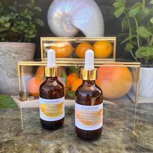 May include: Two amber glass bottles with gold lids and droppers. The bottles are labeled "Follow the Sun" and contain "Golden Facial Oil with Marula Oil" and "Hyaluronic Acid Face Serum with Reishi Mushroom and Marine Collagen Extract".